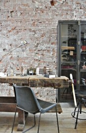 an eclectic home office with an exposed brick accent wall, a wood and glass storage unit, a rough wood desk that feels shabby and industrial and leather chairs