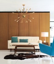 a simple and neutral living room with a plywood covered wall, a sunburst chandelier, a creamy sofa and a blue chair plus matching pillows