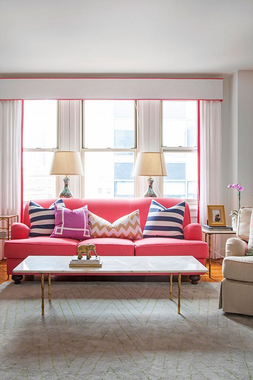 a colorful living room with a pink sofa, pink edging and curtains, a printed rug and cozy furniture
