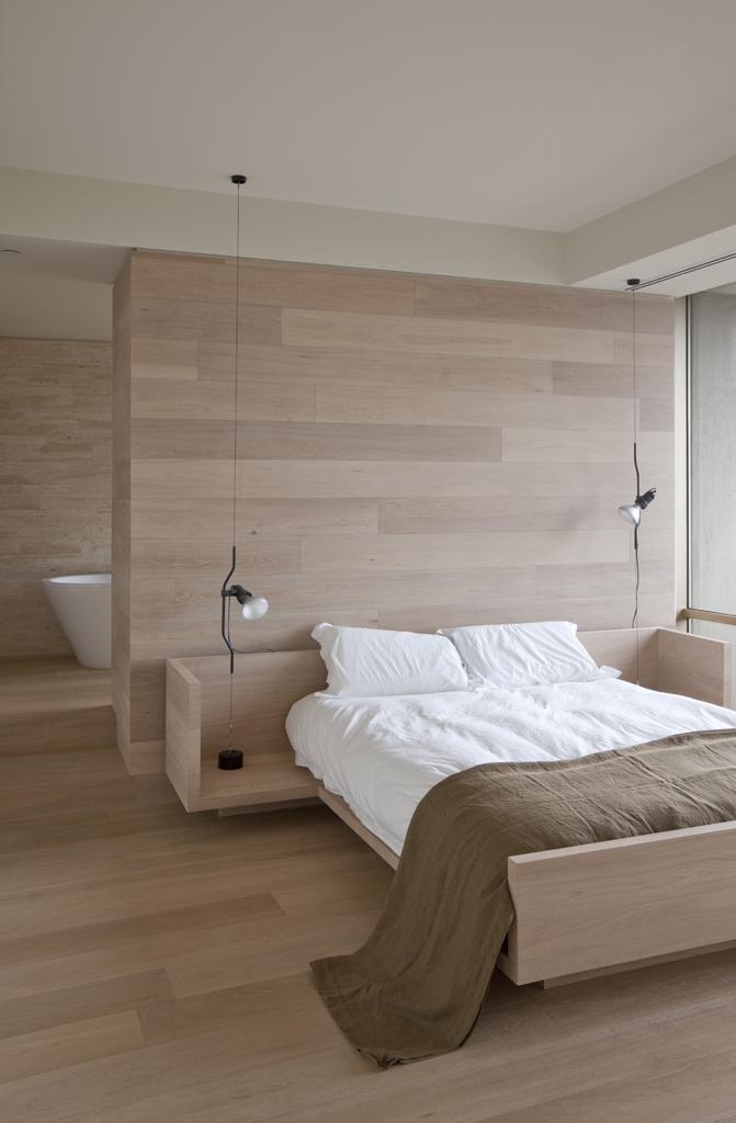A natural minimalist bedroom with a glazed wall, a space divider and a bathroom behind the wall, a light stained bed and neutral bedding