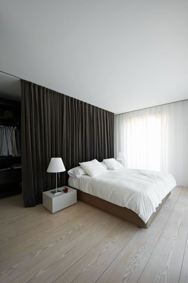 A minimalist bedroom with a walk in closet hidden with a large dark curtain and a neutral bed with white bedding, a sleek nightstand with a white lamp and much natural light