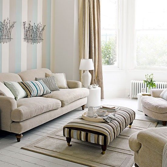 A cozy neutral living room with striped walls, neutral furniture, a striped ottoman and a striped rug is a lovely light filled space