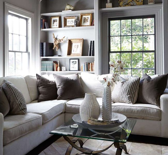 A cozy modern neutral living room with French windows, built in shelves, a sectional and mismatching pillows, a glass coffee table and shiny metallic accents