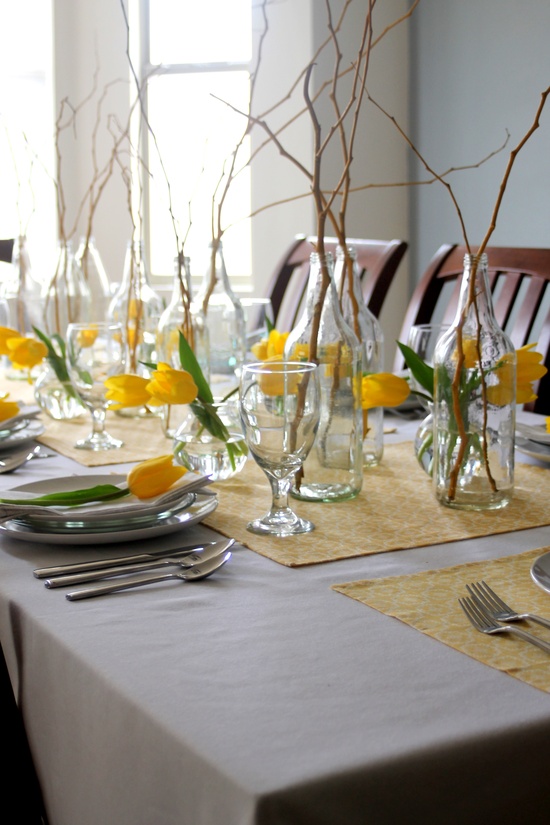 An off white tablecloth, yellow printed placemats, yellow tulips and branches for centerpieces