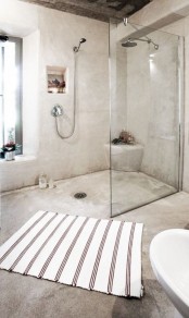 a fully concrete bathroom with a shower space divided with a glass partition, a striped rug and a rough wood ceiling