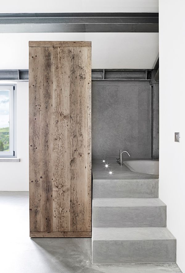 A concrete minimalist bathroom with concrete walls and a floor, a concete staircase and a built in bathtub and a wooden dresser