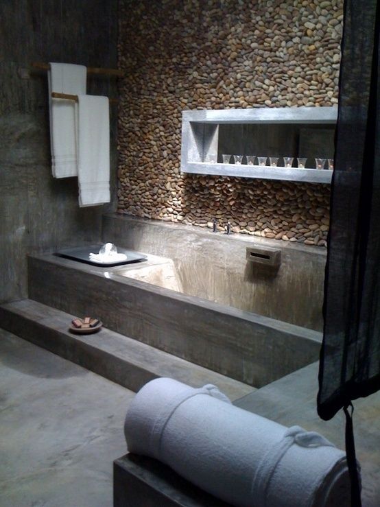 A wabi sabi bathroom made of concrete, with a pebble wall, a built in shelf and a bench, with neutral textiles is cool