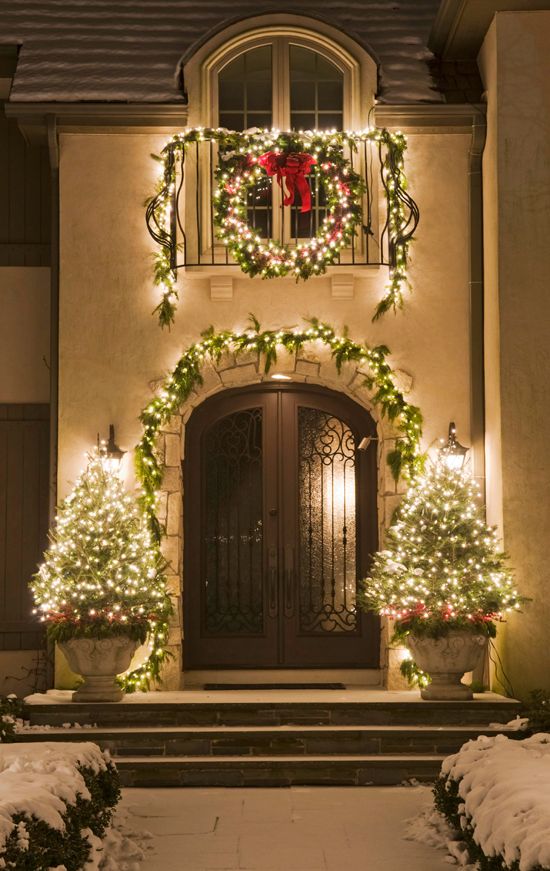 large lit up Christmas trees in urns, an evergreen garland with lights, a matching wreath with a red bow for a Christmas-liek entrance