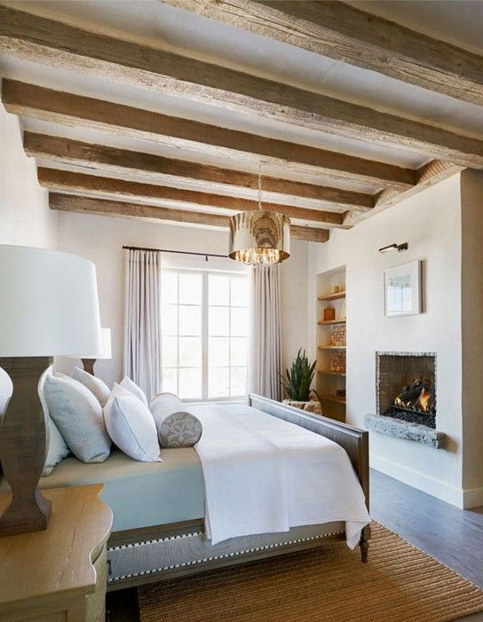 A stylish and chic bedroom with wooden beams, a bed with neutral bedding, a fireplace and built in shelves plus lamps