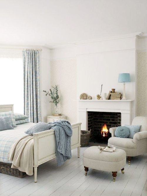 a neutral coastal bedroom with a white fireplace, a bed, a chair and a pouf and delicate light blue accents for a coastal feel