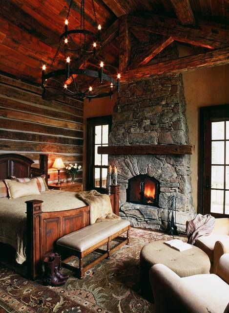 a cozy chalet bedroom with wood clad walls, a stone fireplace, stained furniture, a chandelier and neutral seating furniture