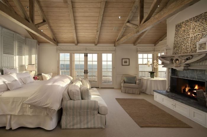 A neutral bedroom with a sea view, a marble clad fireplace, a bed with neutral bedding and a striped sofa for a coastal feel in the space