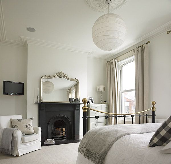 A neutral Parisian bedroom with a fireplace and a mirror, a metal bed with neutral bedding, a white chair and a pendant lamp plus a dresser in the corner