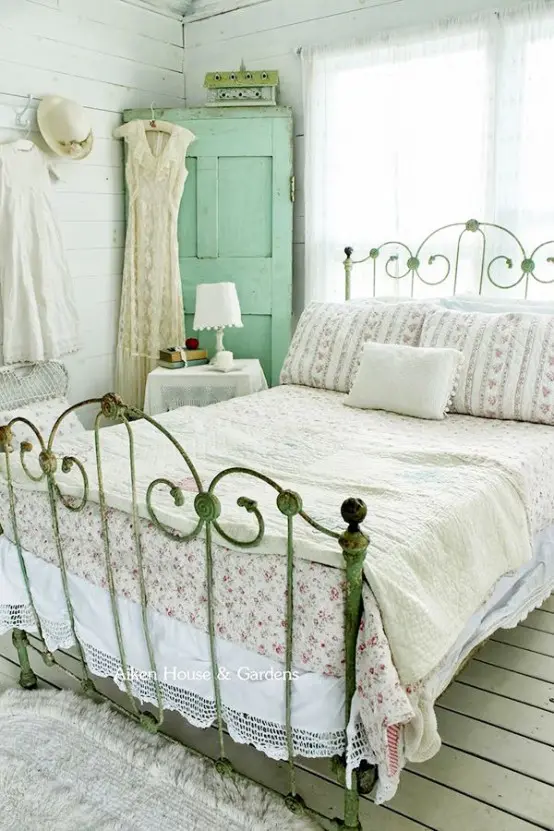 a neutral shabby chic bedroom with a simple wooden floors and walls, a forged bed, a mint storage unit, floral and lace bedding