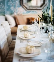 a chic and refined white Thanksgiving tablescape with white linens, porcelain, pumpkins and candles and greenery in bowls is chic