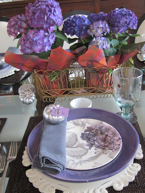 A vintage inspired purple Thanksgiving table setting with purple blooms and plates, brown placemats, silver pumpkins and refined white chargers