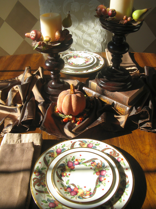 A vintage inspired Thanksgiving tablescape with large and tall candleholders, with faux apples and pumpkins, printed plates and neutral linens is a chic and cool idea