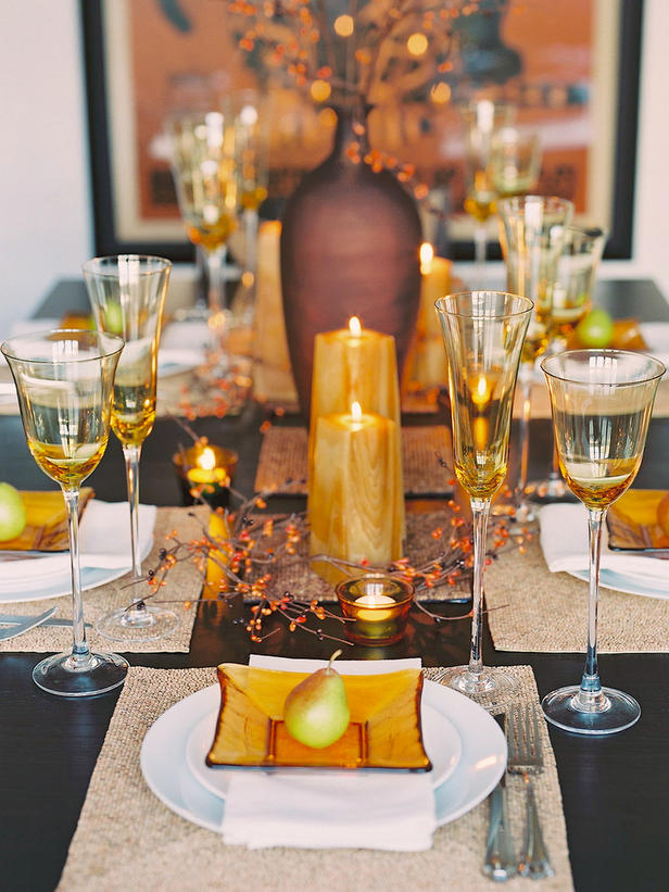 A chic warm colored Thanksgiving table setting with amber glasses and plates, with pillar candles, white porcelain and woven placemats plus faux branches