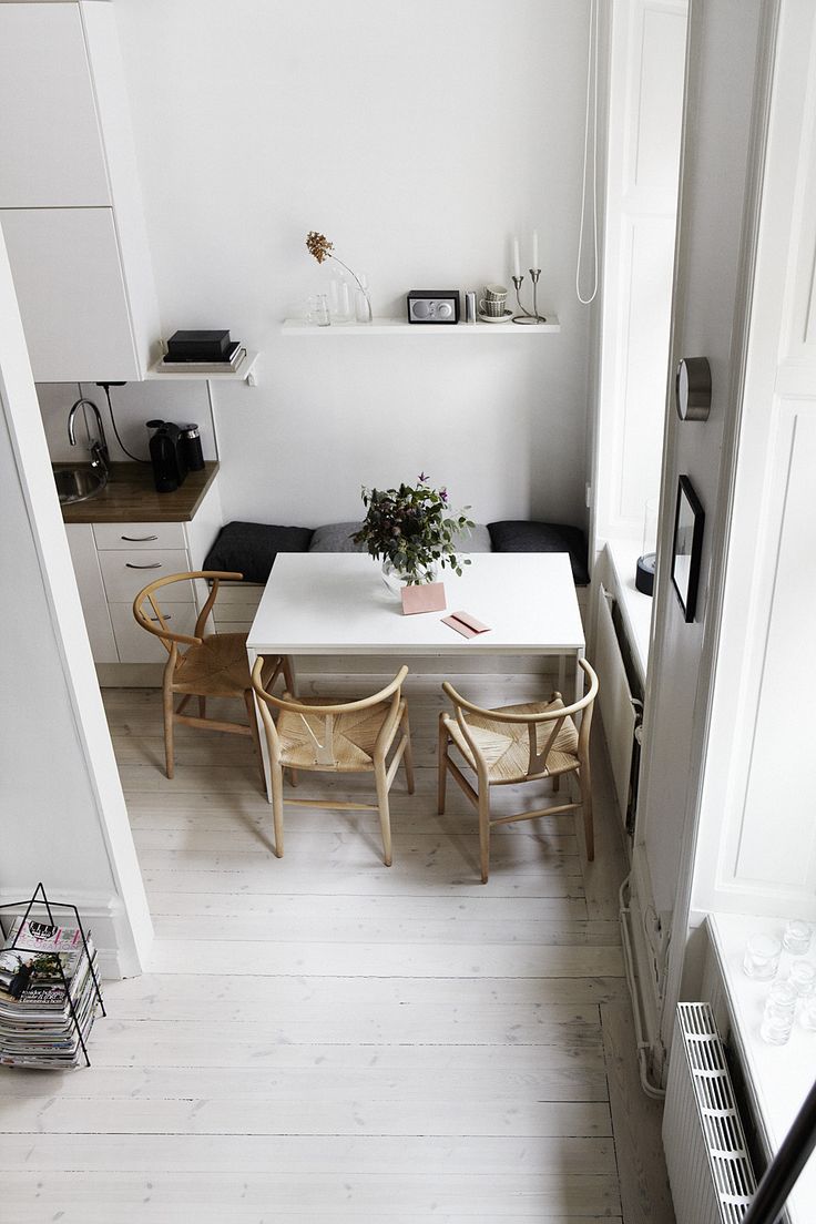 A small yet functional Scandinavian dining space with a bench with cushions, woven chairs, a built in bench and a white table plus an open shelf over the space