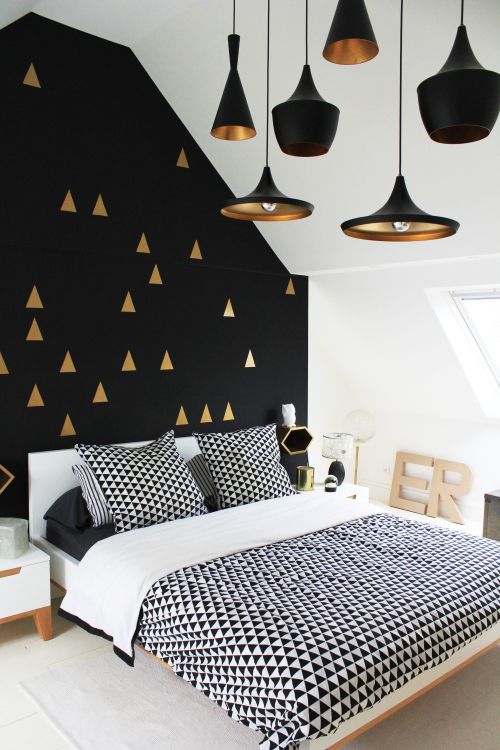 a modern and contrasting bedroom with a black and gold triangle accent wall, neutral furniture, black and white geometric print bedding and gold and black pendant lamps