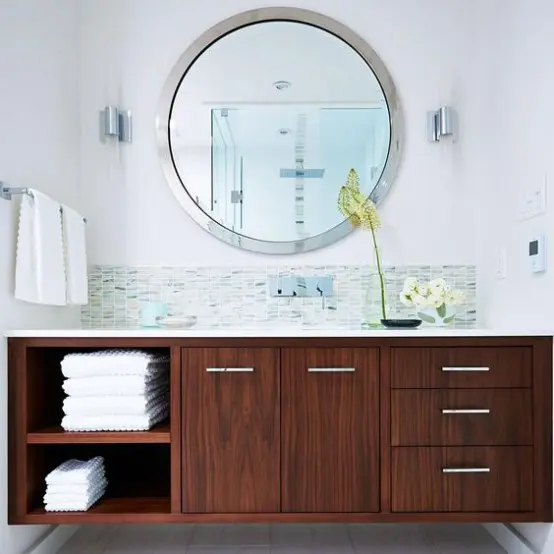 a neutral bathroom with a grey and green tile backsplash and a rich stained vanity plus a round mirror and potted blooms
