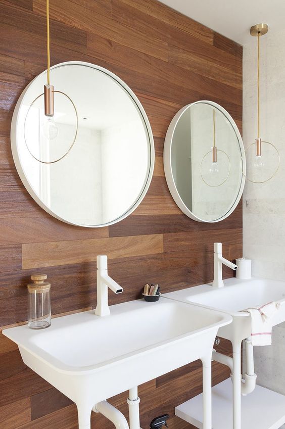 A chic mid century modern bathroom with a wooden accent wall, white appliances and catchy pendant lamps over the sinks