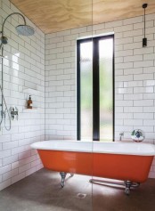 a chic mid-century modern bathroom with white tiles, a plywood ceiling, a bold orange clawfoot tub for a colorful accent