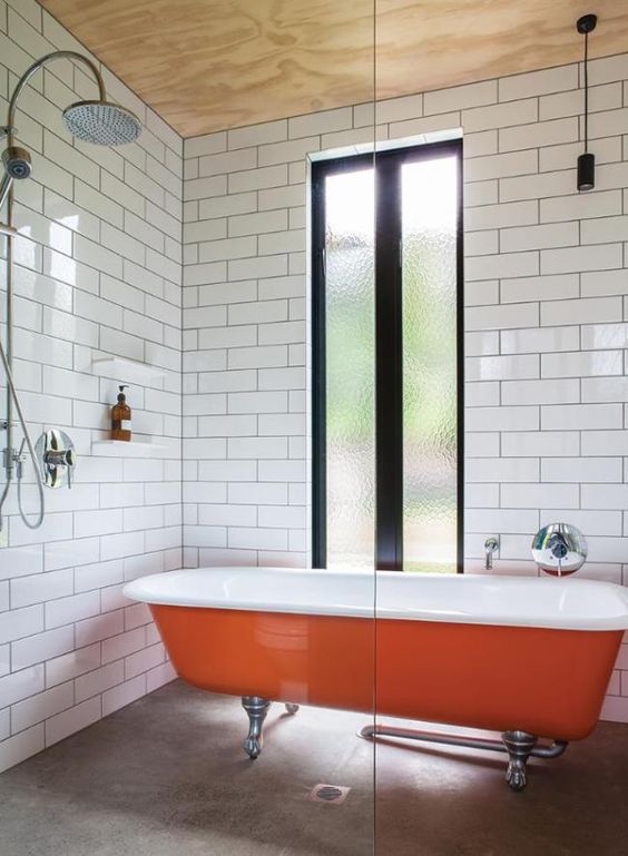 A chic mid century modern bathroom with white tiles, a plywood ceiling, a bold orange clawfoot tub for a colorful accent