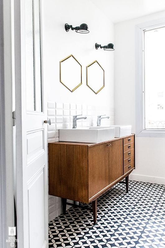 An elegant mid century modern bathroom with black and white tiles on the floor, a mid century modern vanity of wood, hex mirrors in gold frames