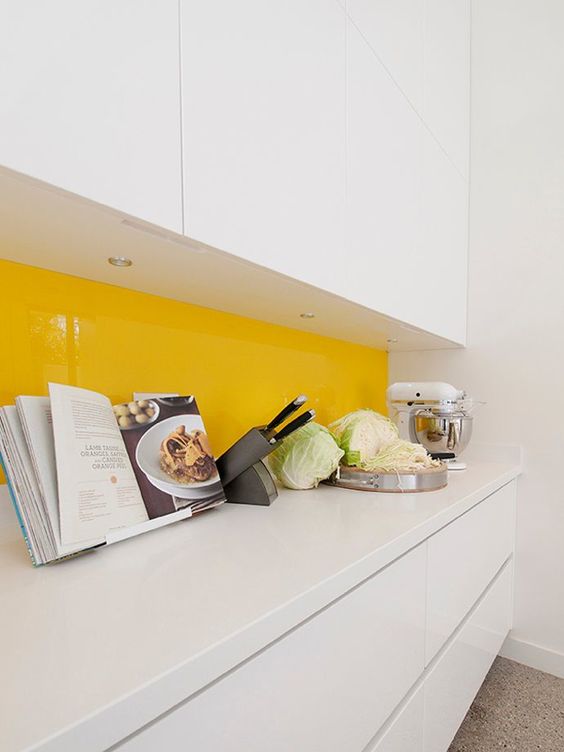 a minimalist white kitchen with white countertops and a yellow solid glass backsplash that brings a juicy touch of color and gloss