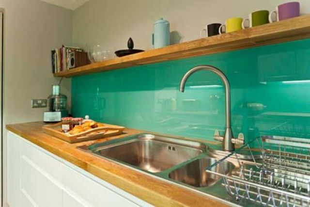 A modern white kitchen with light stained butcherblock countertops and a bold green glass backsplash that contrasts the cabinets