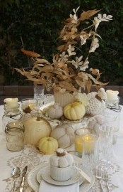 a vintage rustic Thanksgiving tablescape with a lace runner, white textiles, a dried leaf and flower centerpiece, white and neutral pumpkins, candles in glasses and white porcelain is a chic one