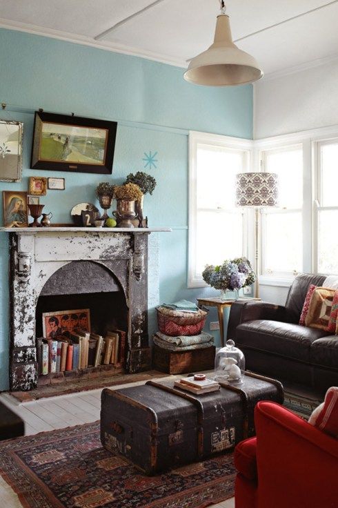 A vintage meets eclectic living room with a blue accent wall, a non working fireplace used for book storage, a gallery wlal and a pendant lamp plus a chest as a coffee table