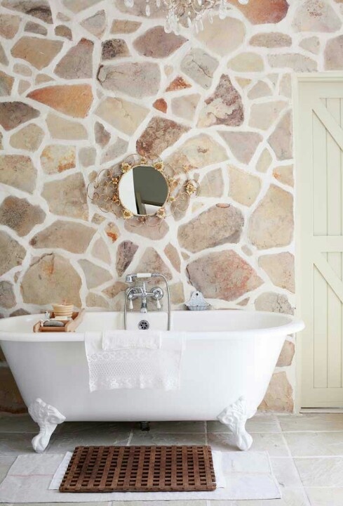 A light filled farmhouse bathroom with a natural stone wall, a refined mirror and a clawfoot bathtub