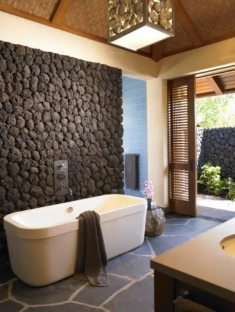 An outdoor indoor contemporary bathroom with a stone floor and a decorative stone accent wall to make the bathtub stand out