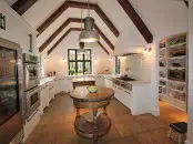 a white kitchen with shaker style cabinets, white stone countertops and dark beams on the ceiling, a round stained kitchen island that is a small table at the same time