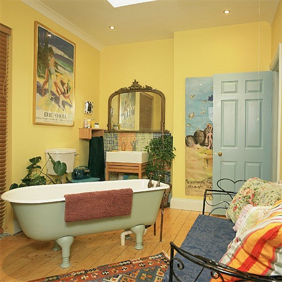 a whimsical yellow bathroom with a green clawfoot bathtub, a large mirror, retro artwork, a wall mural and a metal daybed with bright bedding